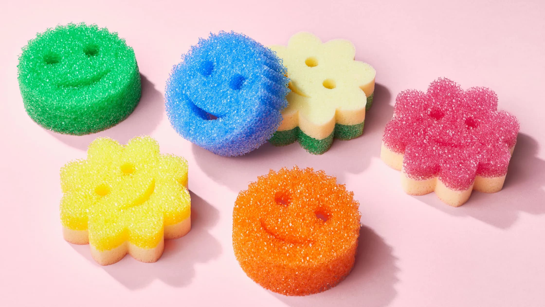 Six different Scrub Daddy colourful sponges are placed over a pale pink surface.