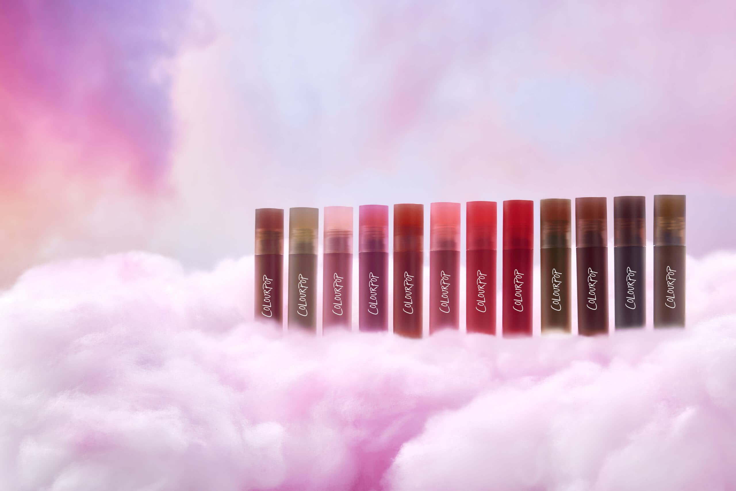 Twelve shades of ColourPop CloudWhip Lipstick are aligned on a cloudy pink environment.