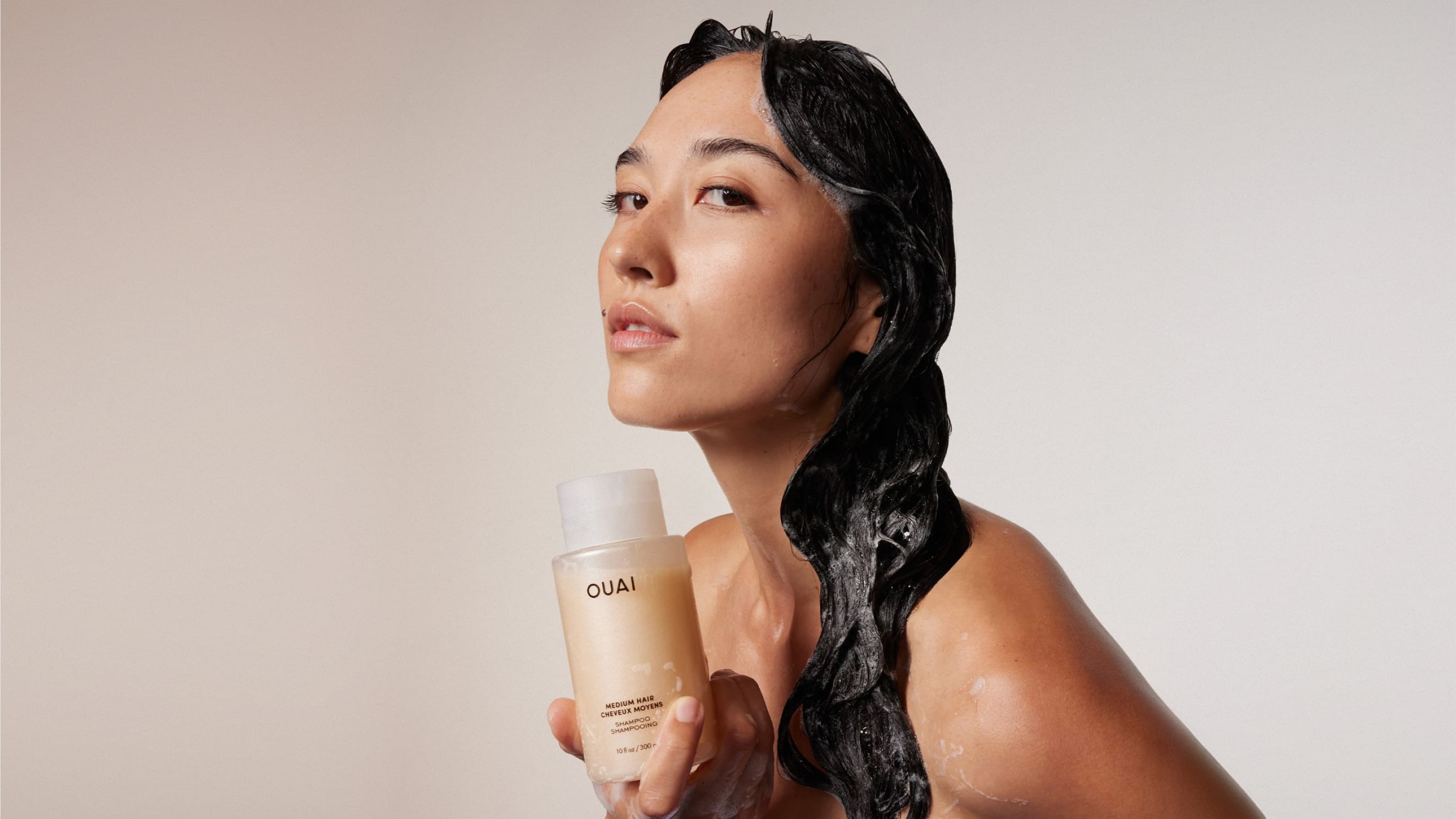 A person with long dark and wet hair is holding a bottle of OUAI shampoo over a soft warm beige coloured background