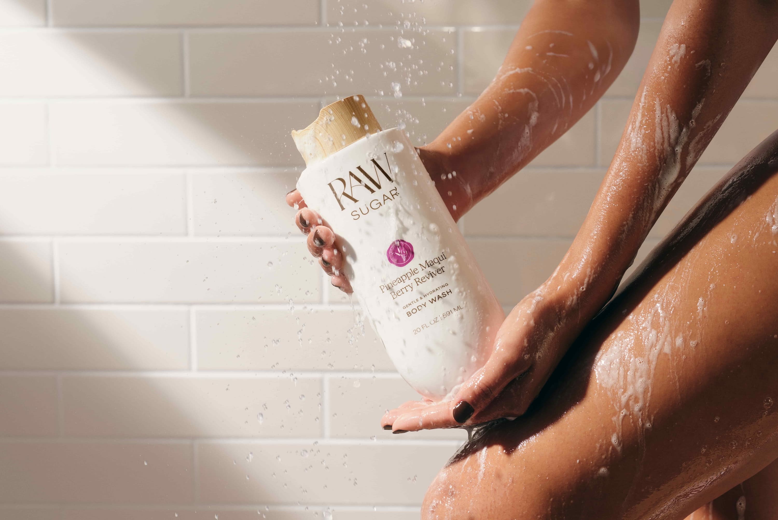 A person with soap foam on their skin is holding a jar of RAW Sugar body wash in front of white tiles.
