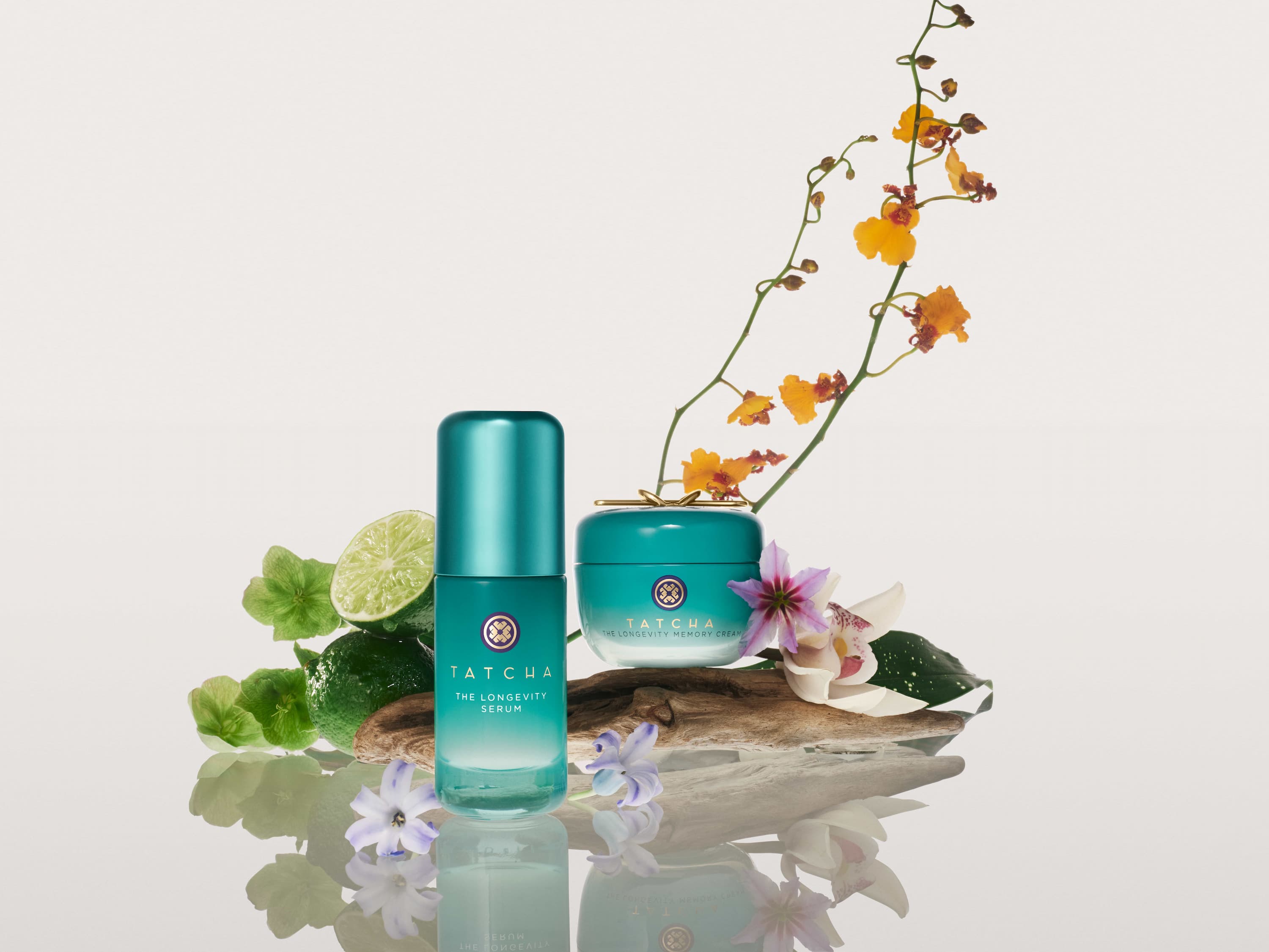 Two Tatcha Longevity serum and cream product are standing on a white reflective surface, surrounded by fruits and delicate flowers.