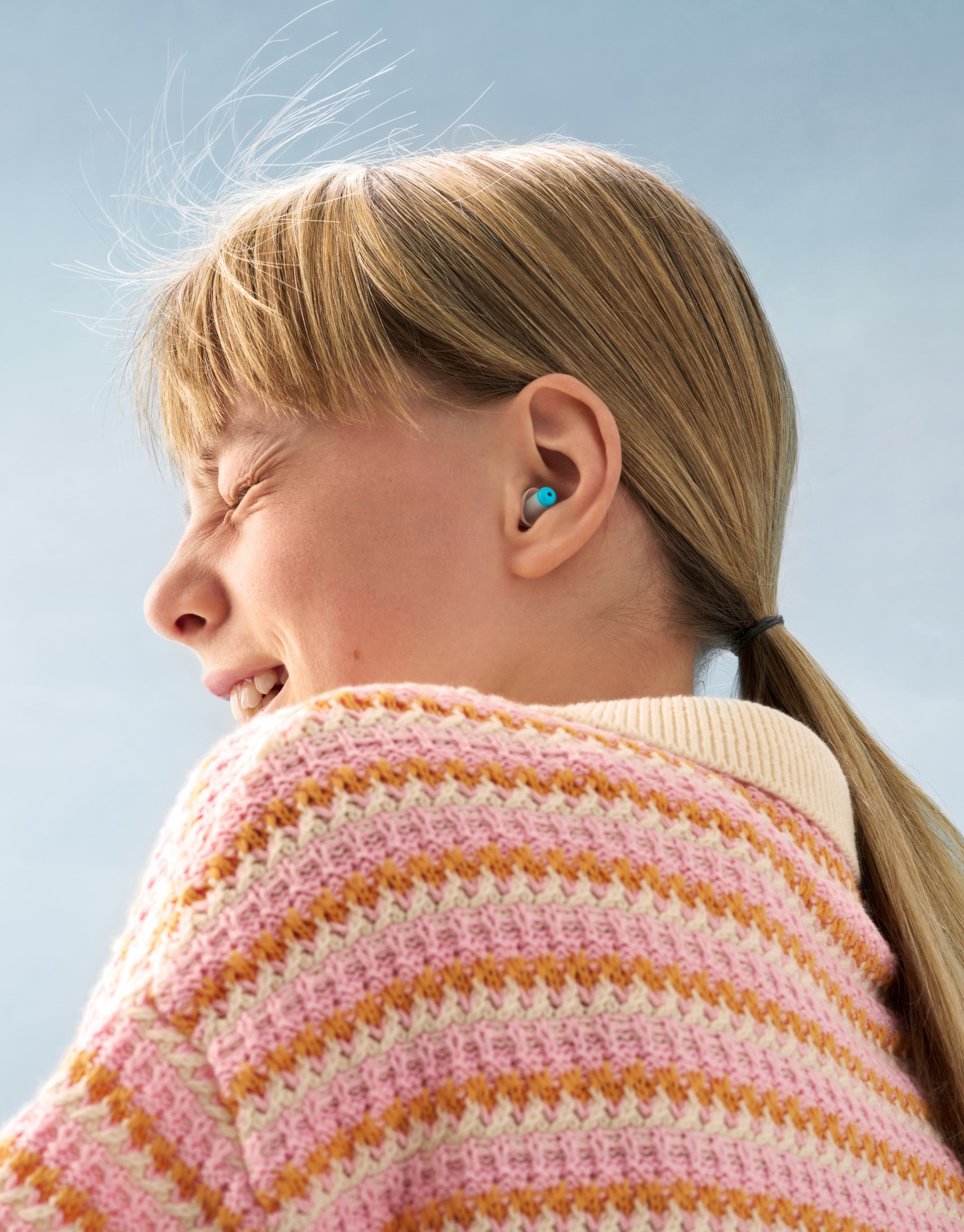 A young blond person is wearing blue earplugs and a knitted colourful sweater while smiling in the wind.
