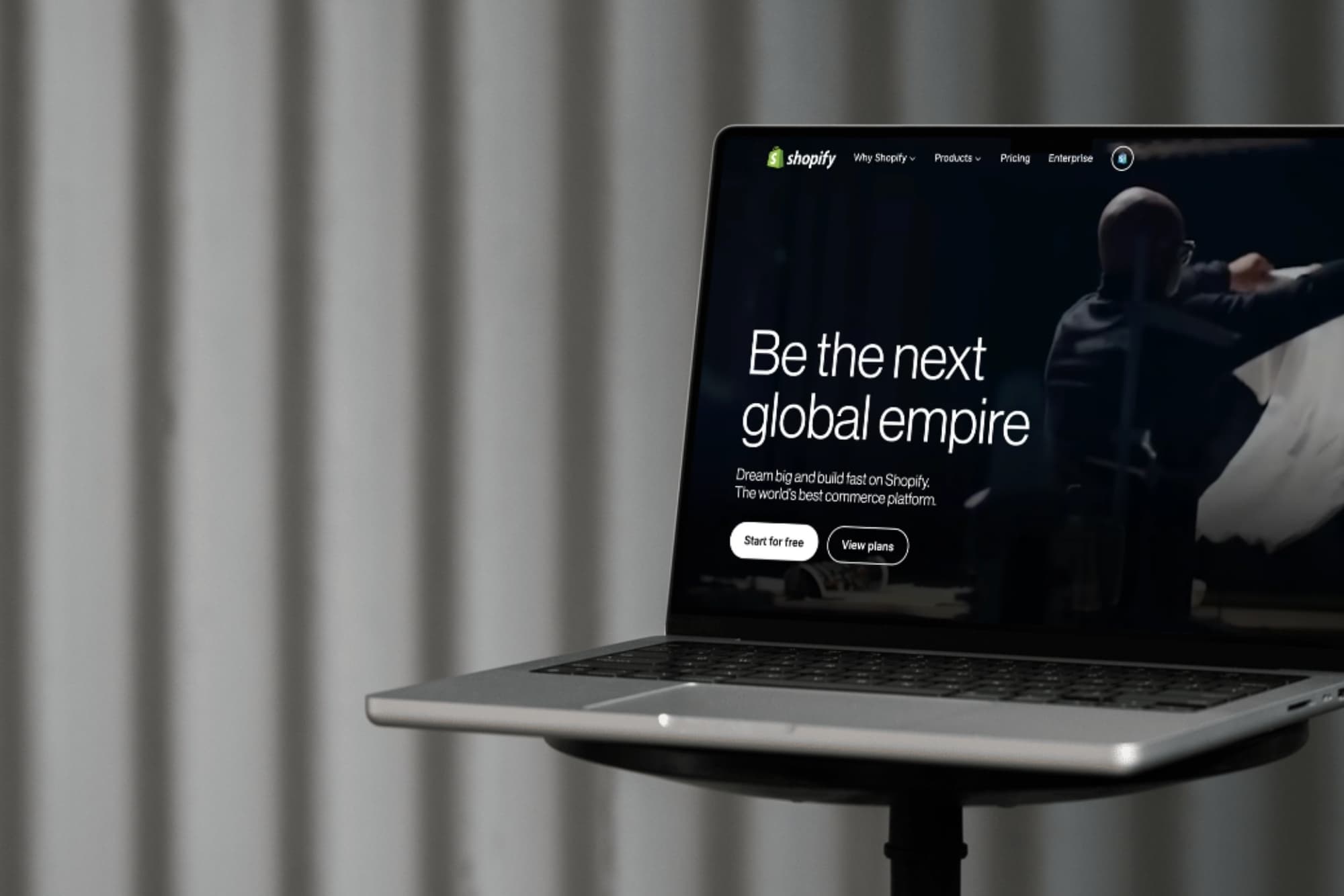 Laptop displaying Shopify's homepage with the message "Be the next global empire" against a blurred curtain background.