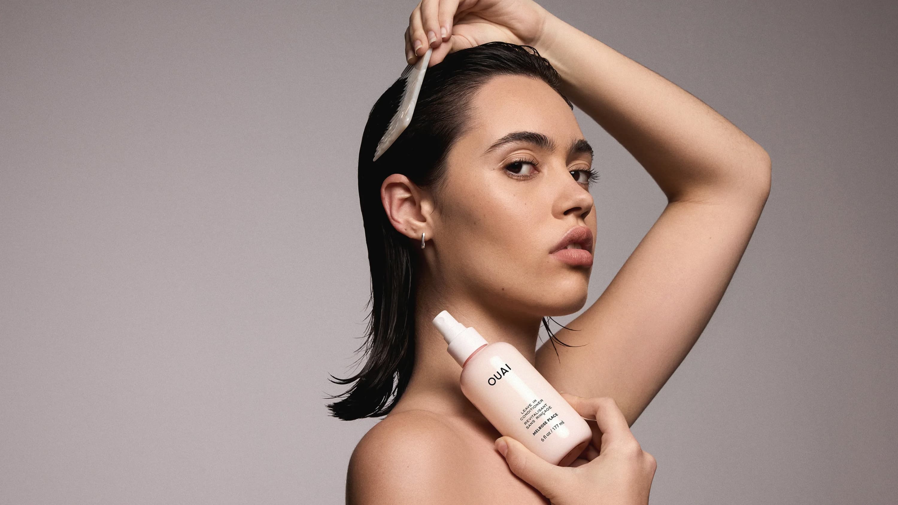A person is brushing their wet dark hair while holding a pale pink bottle of OUAI