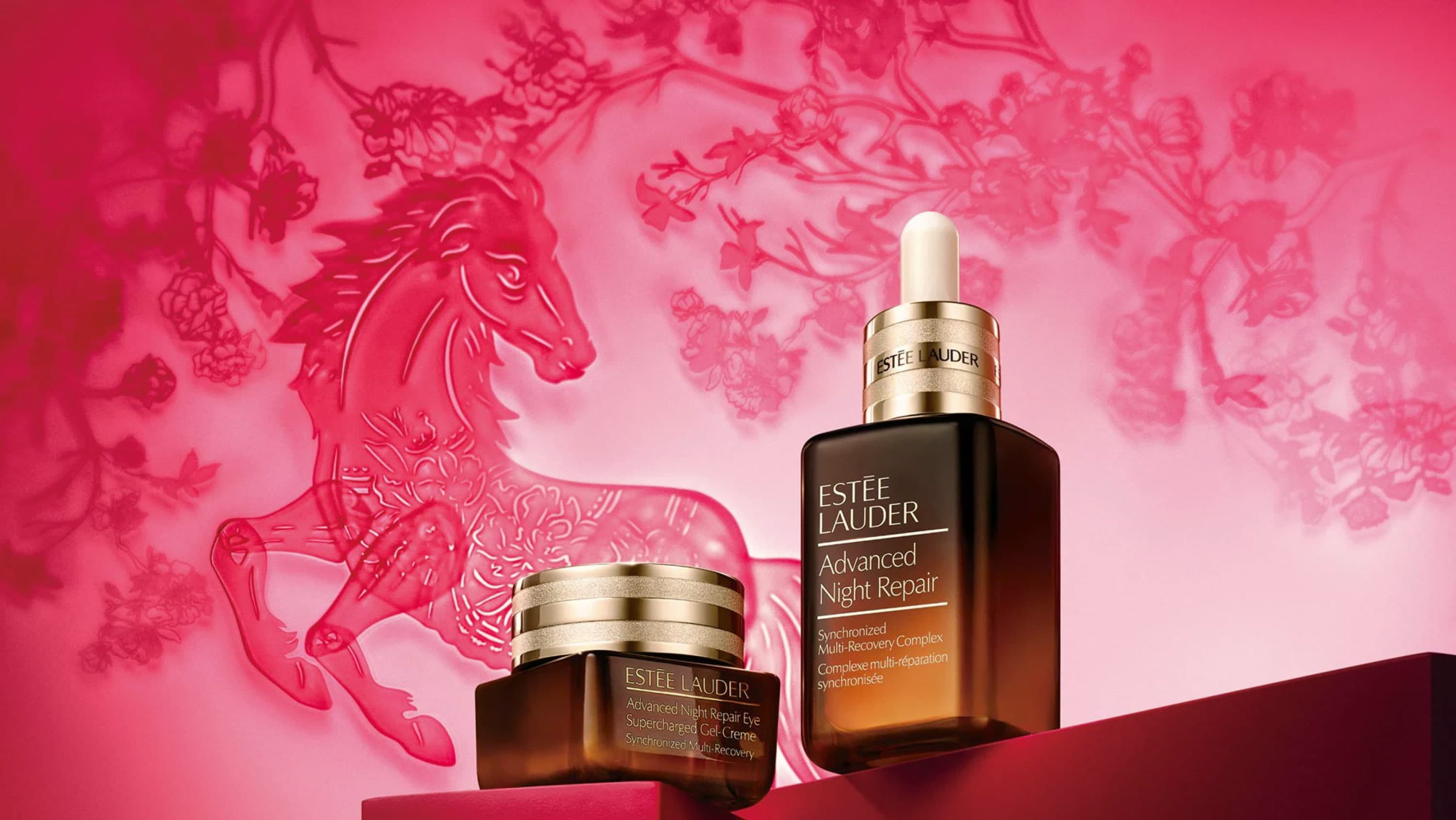 Two Estée Lauder Advanced Night Repair products are placed on a deep red surface with an illustration of a red horse in the background surrounded by a flowering tree.