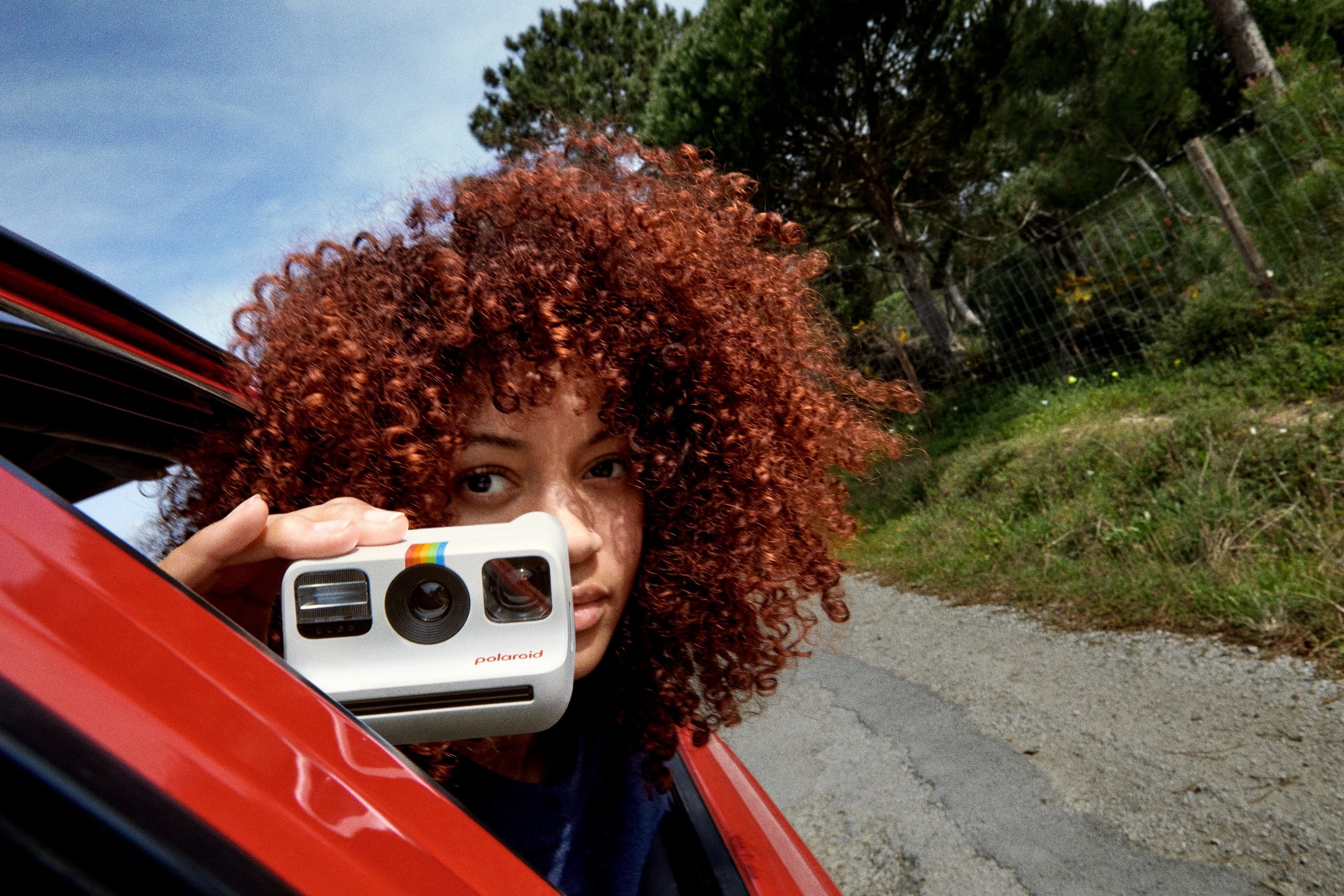 A person with curly hair holding a white Polaroid camera in a red car.