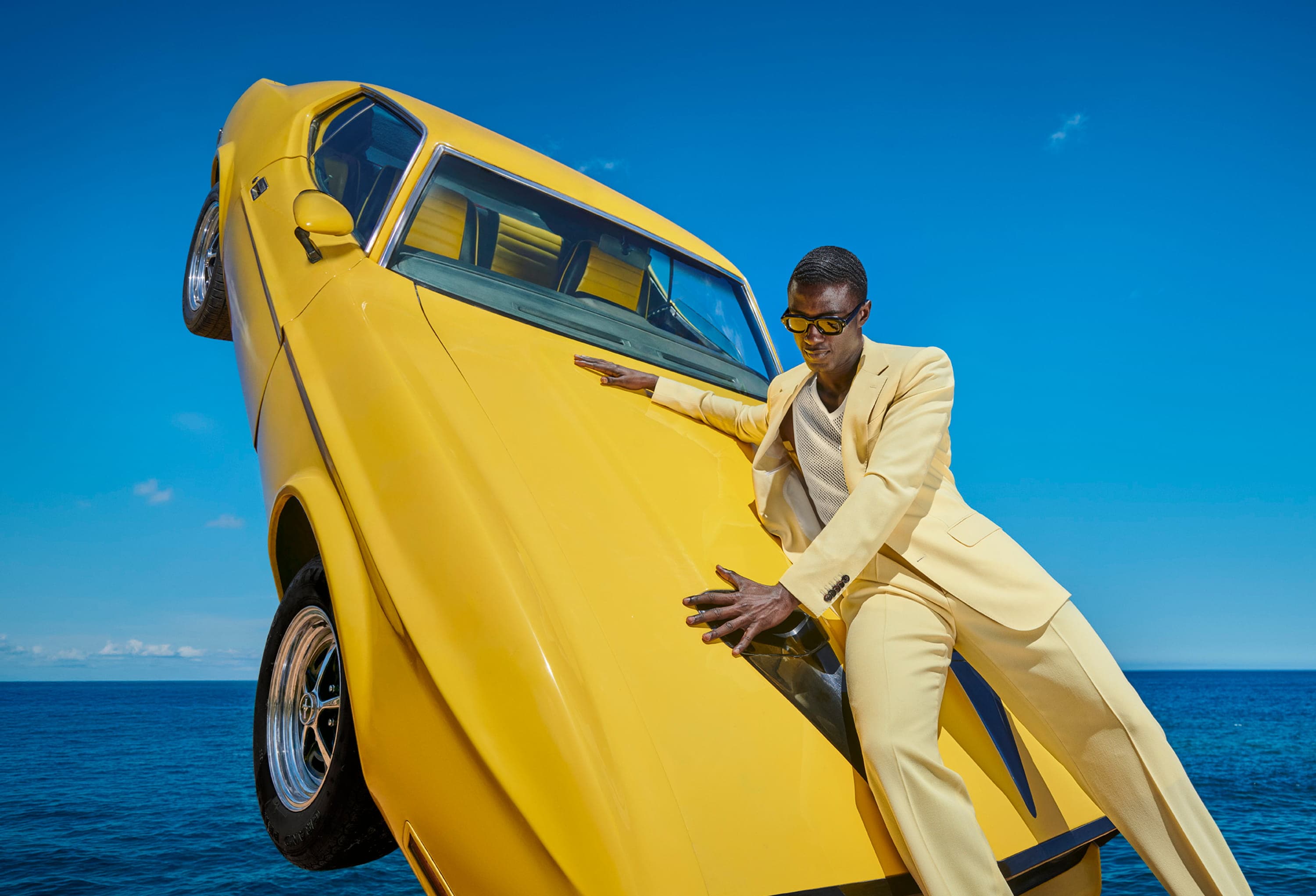 A person in a yellow Suitsupply suit leans against a yellow car, which is angled vertically against a clear blue sky and ocean backdrop.
